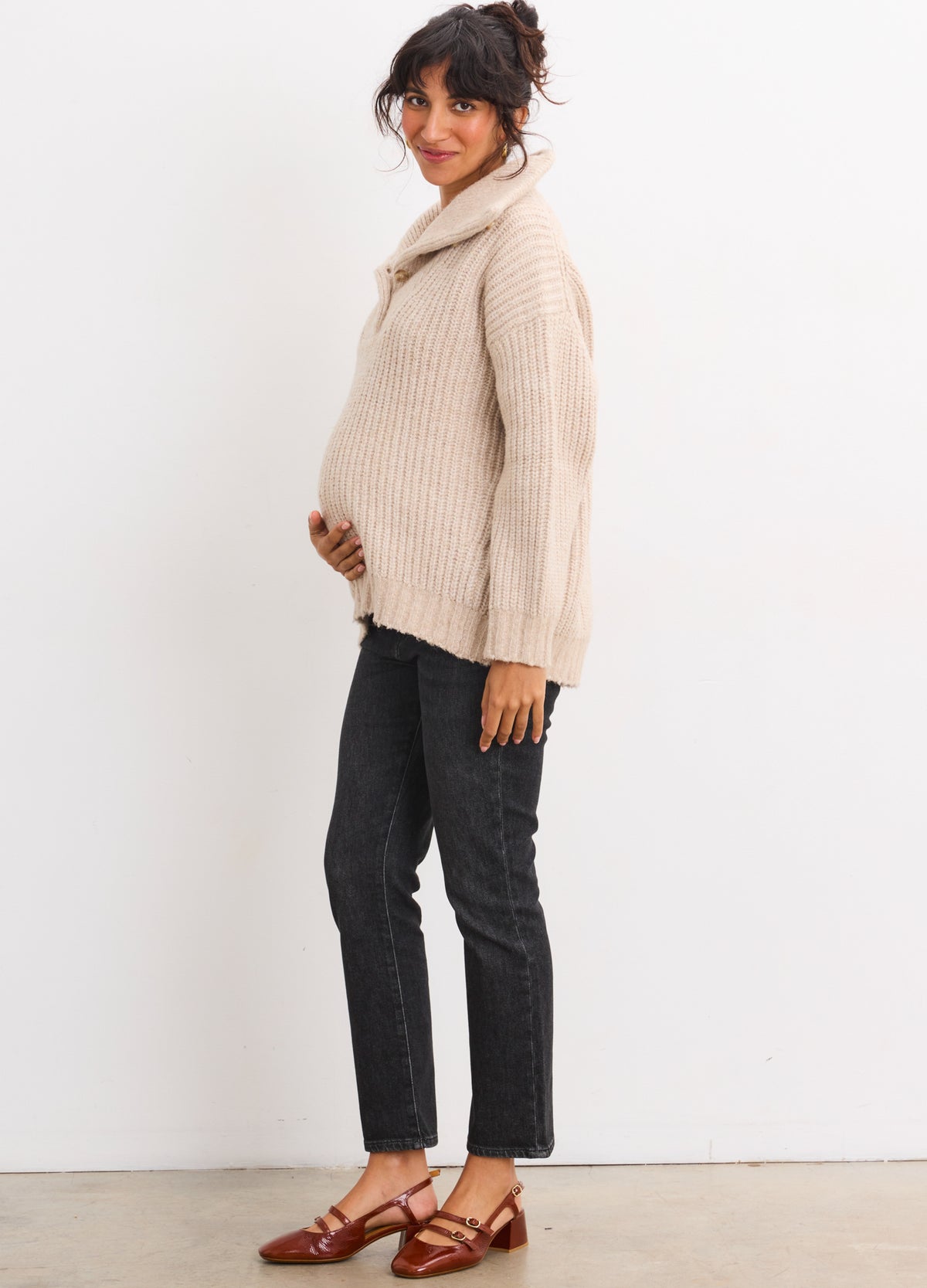 Pregnant woman wearing oatmeal maternity sweater with cozy turtleneck and long sleeves paired with black pants and brown.