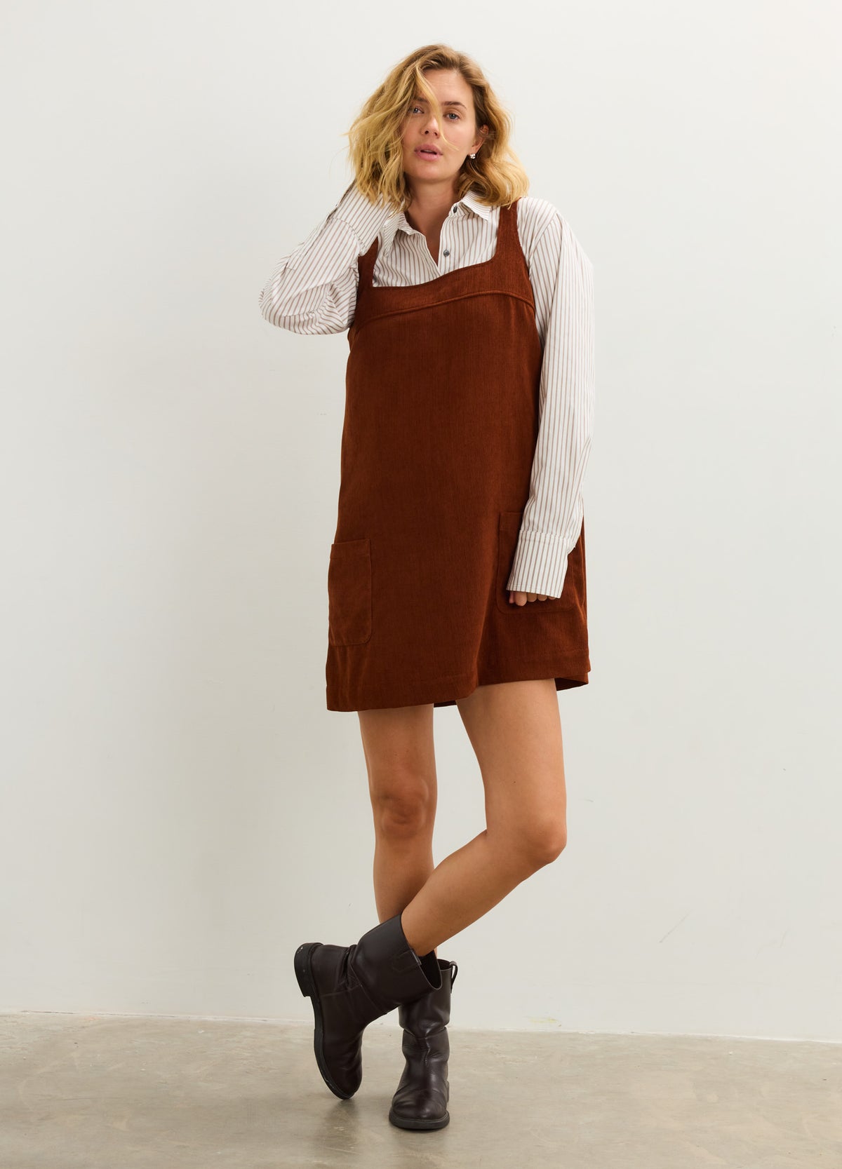 Rust brown corduroy mini dress with adjustable straps, loose maternity fit, worn over white long-sleeve blouse.