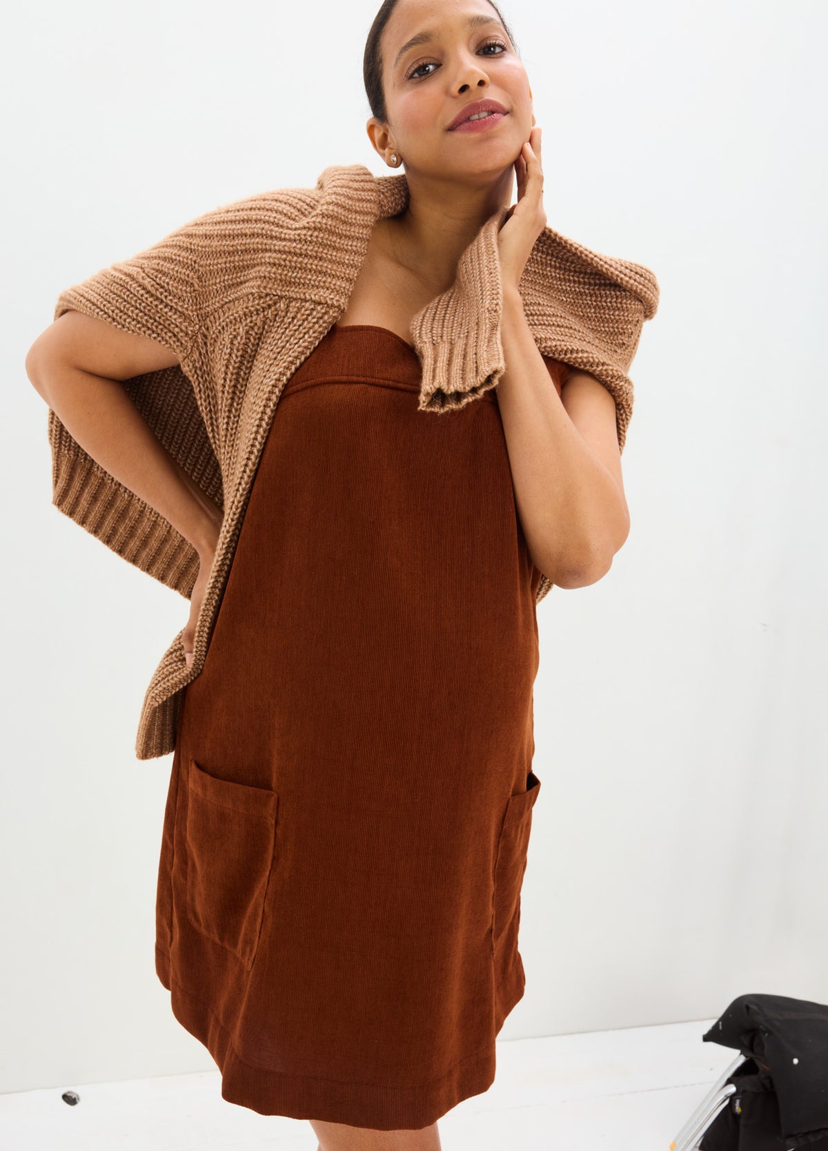 Brown corduroy maternity mini dress with square neckline and front pockets, paired with beige knit shawl.