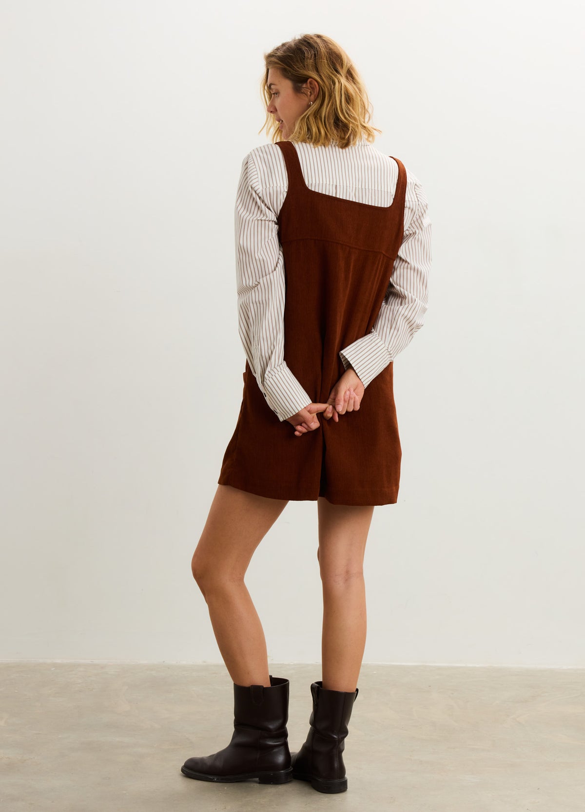 Brown corduroy mini dress with square neckline and wide straps, worn over white long-sleeve shirt, paired with black ankle.