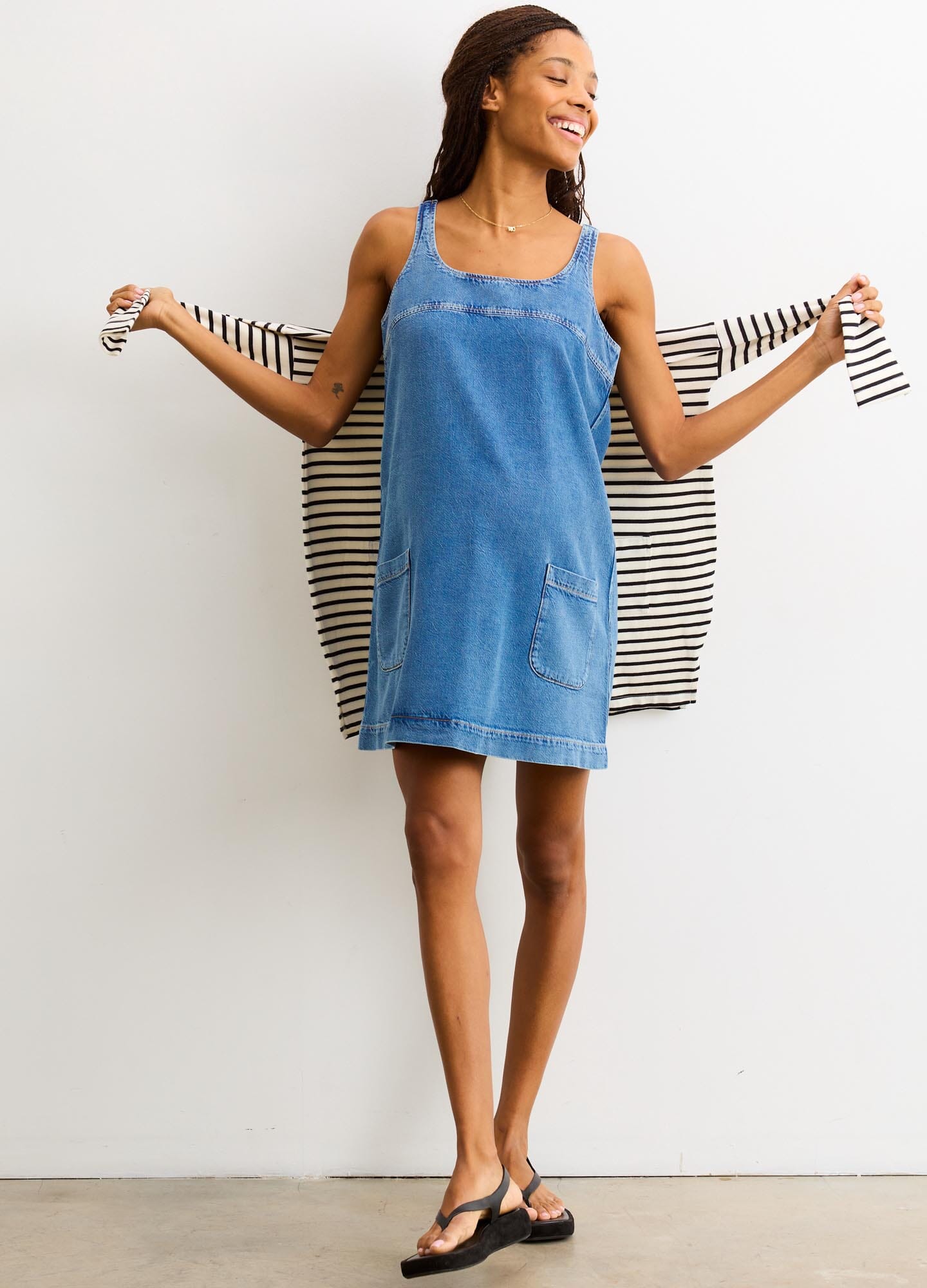 Blue denim mini dress with square neckline and adjustable straps, styled for maternity, worn with striped cardigan draped.
