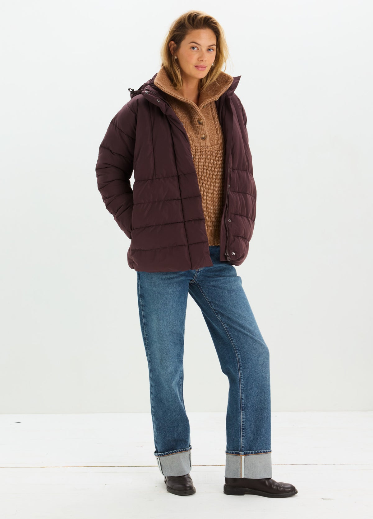 Woman wearing a dark plum quilted puffer jacket with a hood over a tan sweater and jeans.