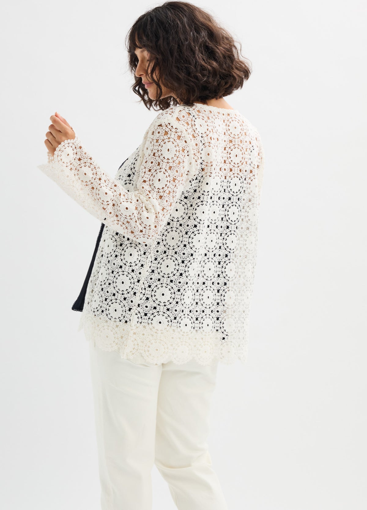 White crochet knit top with floral pattern, long sleeves, and scalloped hem, worn over black maternity tank.