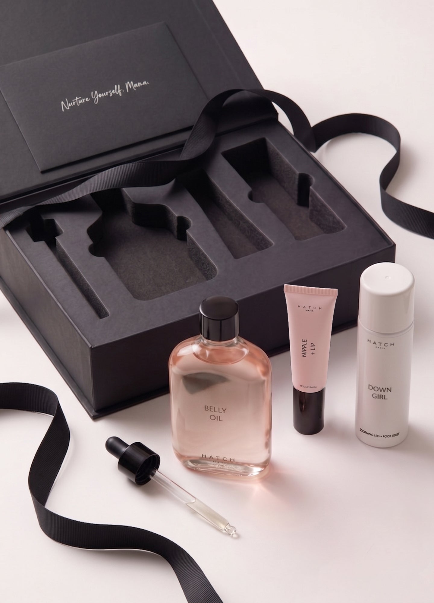 Black gift box with cutouts holding clear pink liquid bottle with dropper, pink cream tube, and white cylindrical bottle,.