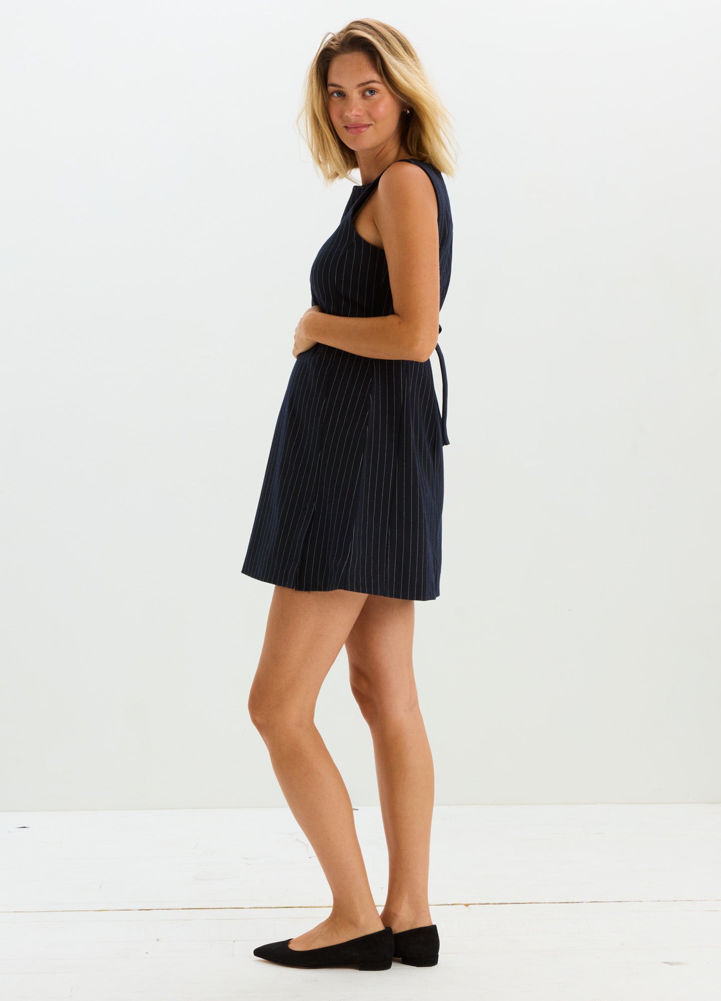 Black sleeveless maternity mini dress with subtle pinstripe pattern and a tied back belt.