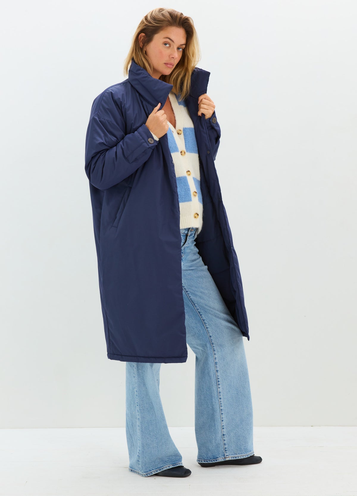 A woman modeling a long navy blue puffer coat over a cream and blue checkered top with light blue wide-leg jeans, standing against a plain white studio background.