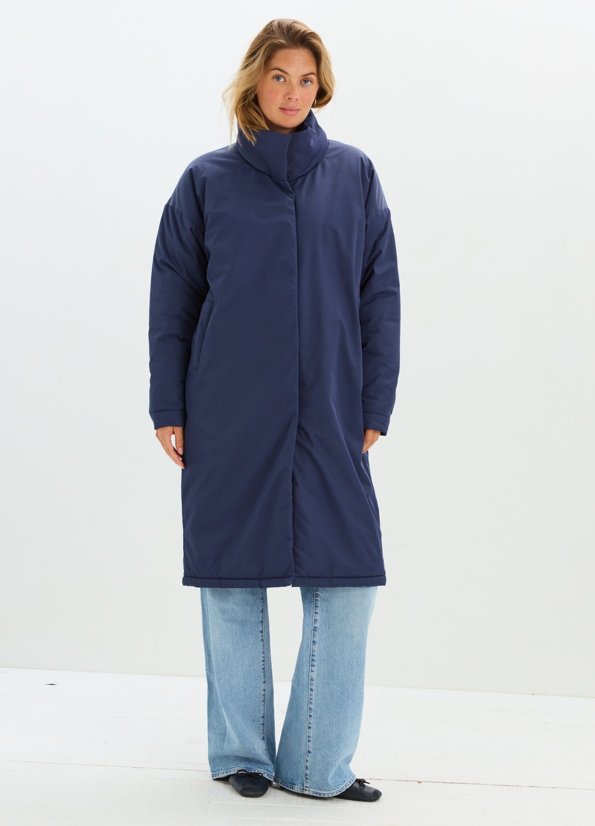 Model wearing a long navy padded coat with a high collar, front zip, and light blue jeans.
