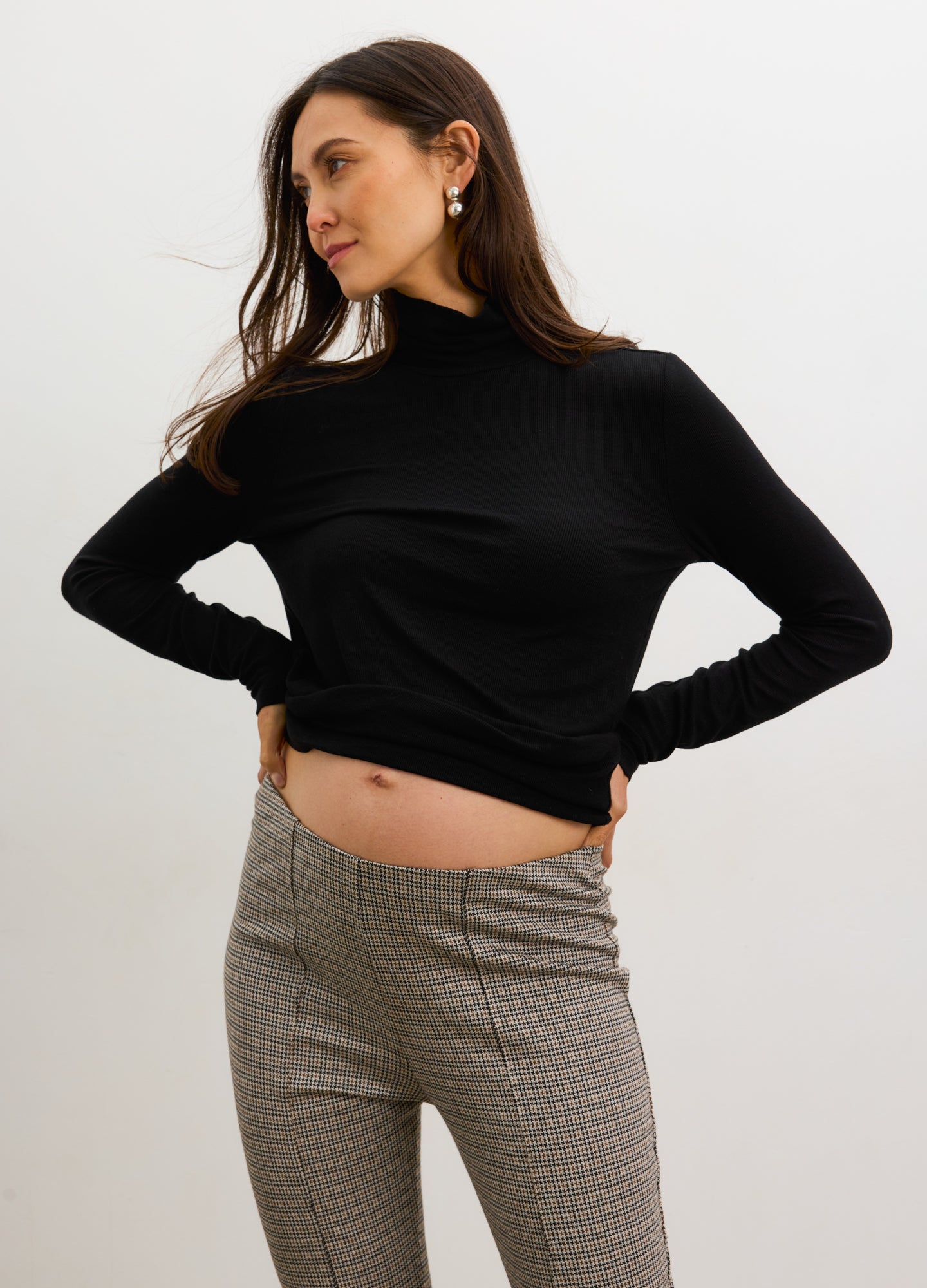 Pregnant woman wearing a black long-sleeve top and gray checkered pants against a white background