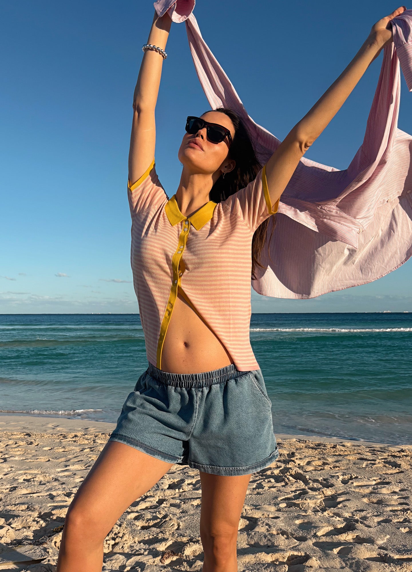 Light pink and yellow striped maternity shirt with buttons and short sleeves, styled open over casual denim shorts on a.