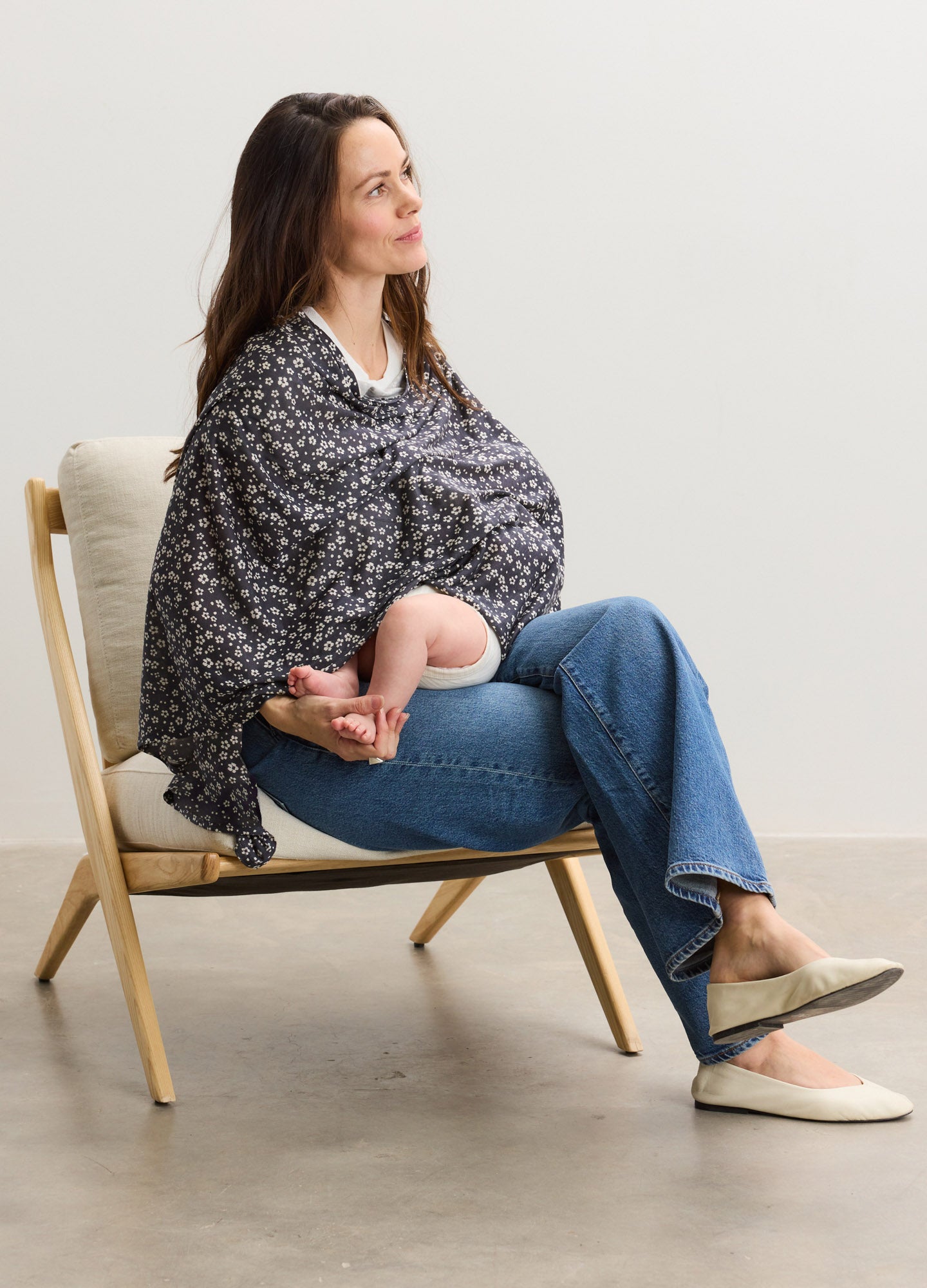 Black and white floral nursing cover draped over a seated woman wearing jeans and slippers, breastfeeding a baby.
