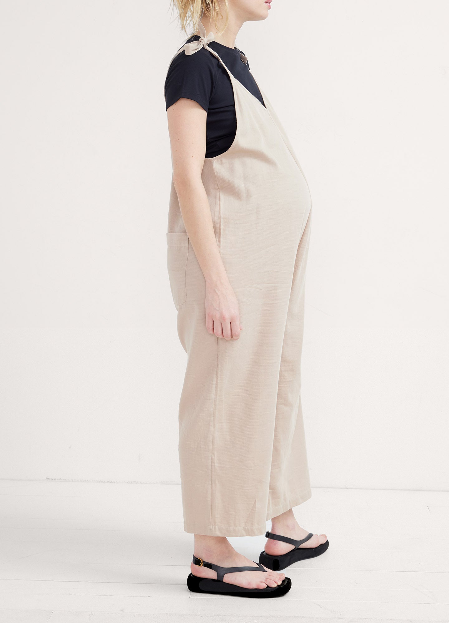 Beige sleeveless maternity jumpsuit with wide legs, worn over a black short-sleeve top, paired with black sandals.