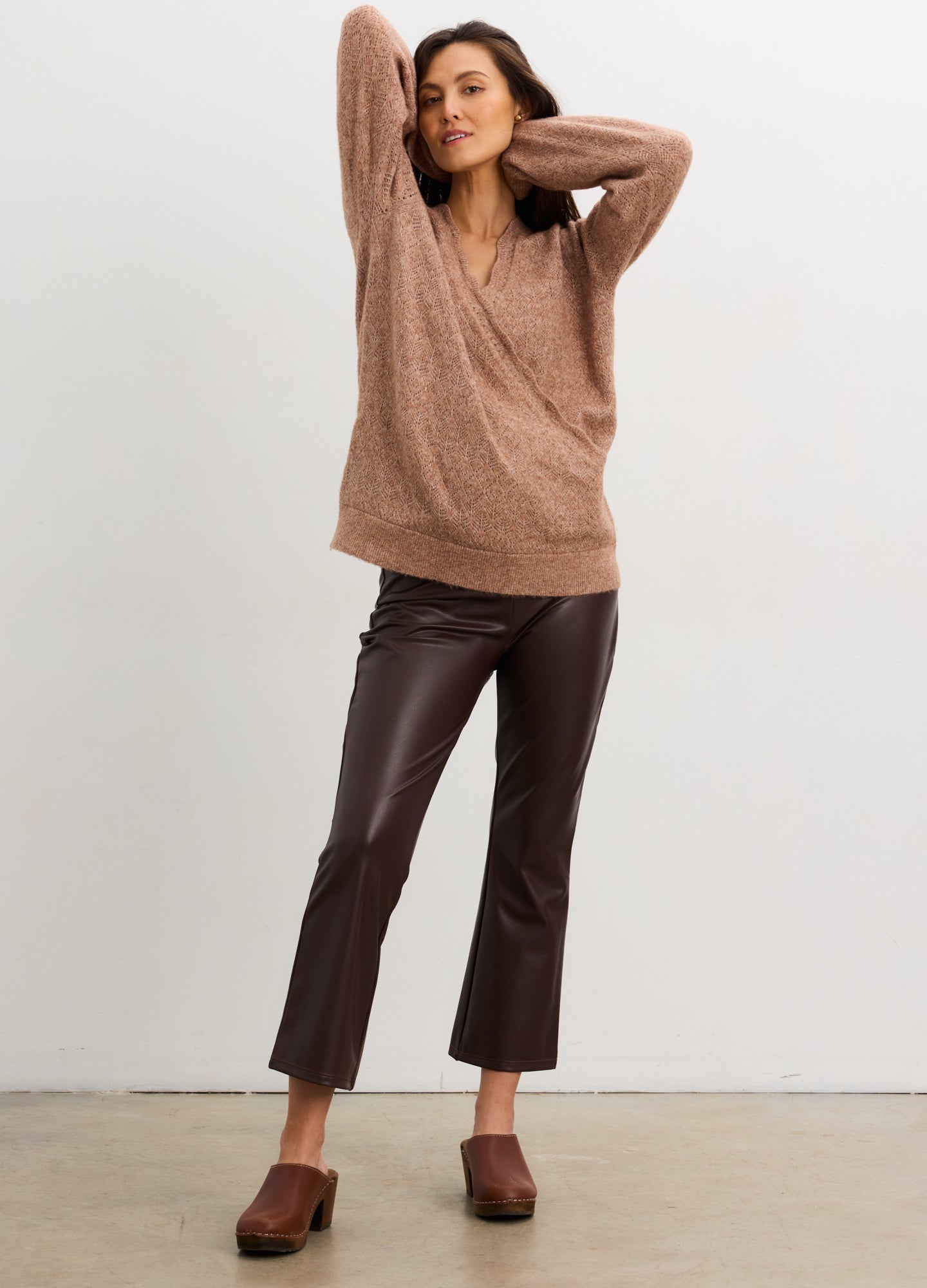 Pregnant woman wearing a brown sweater and leather pants against a white background