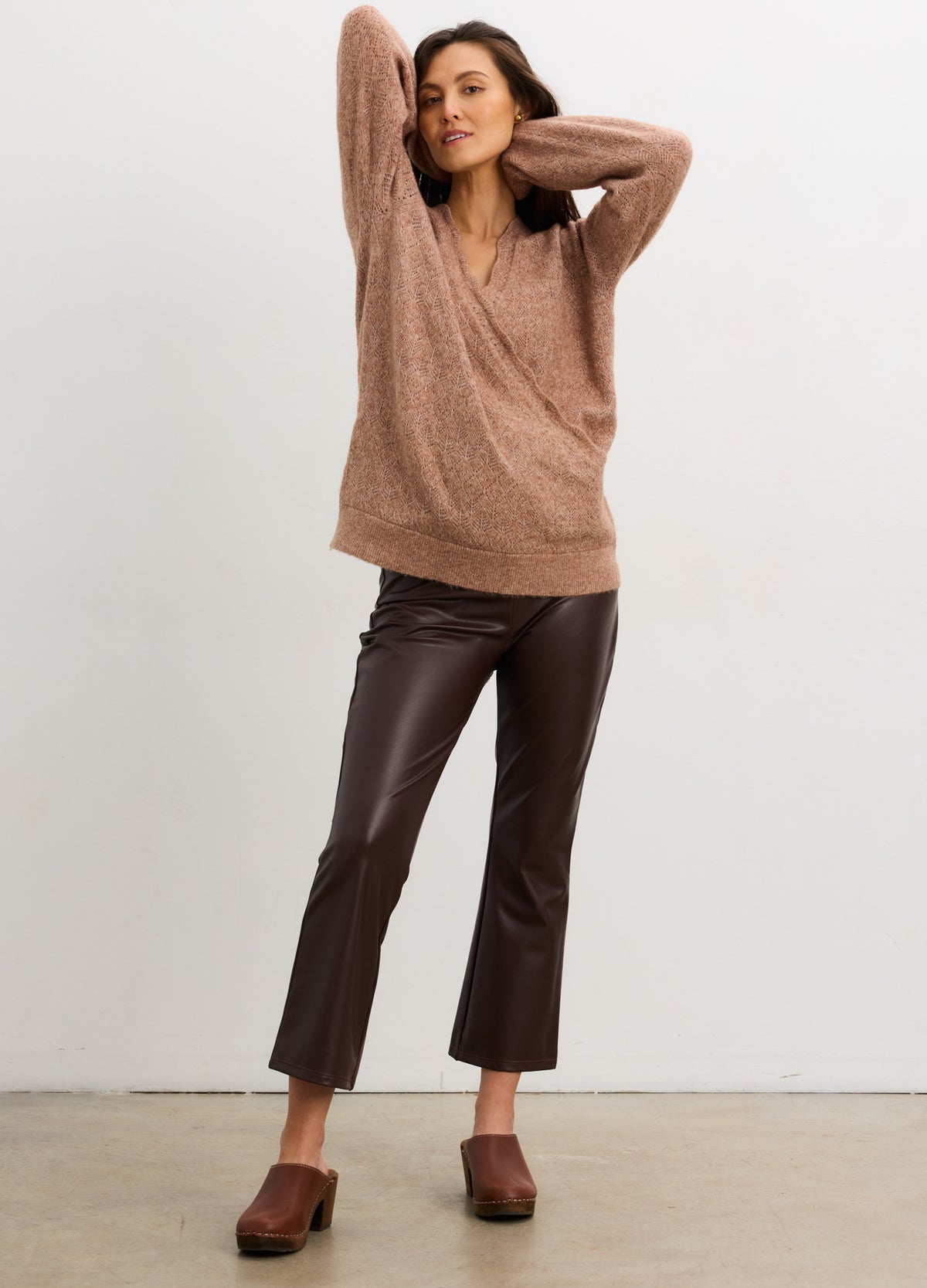 Pregnant woman wearing a brown sweater and leather pants against a white background