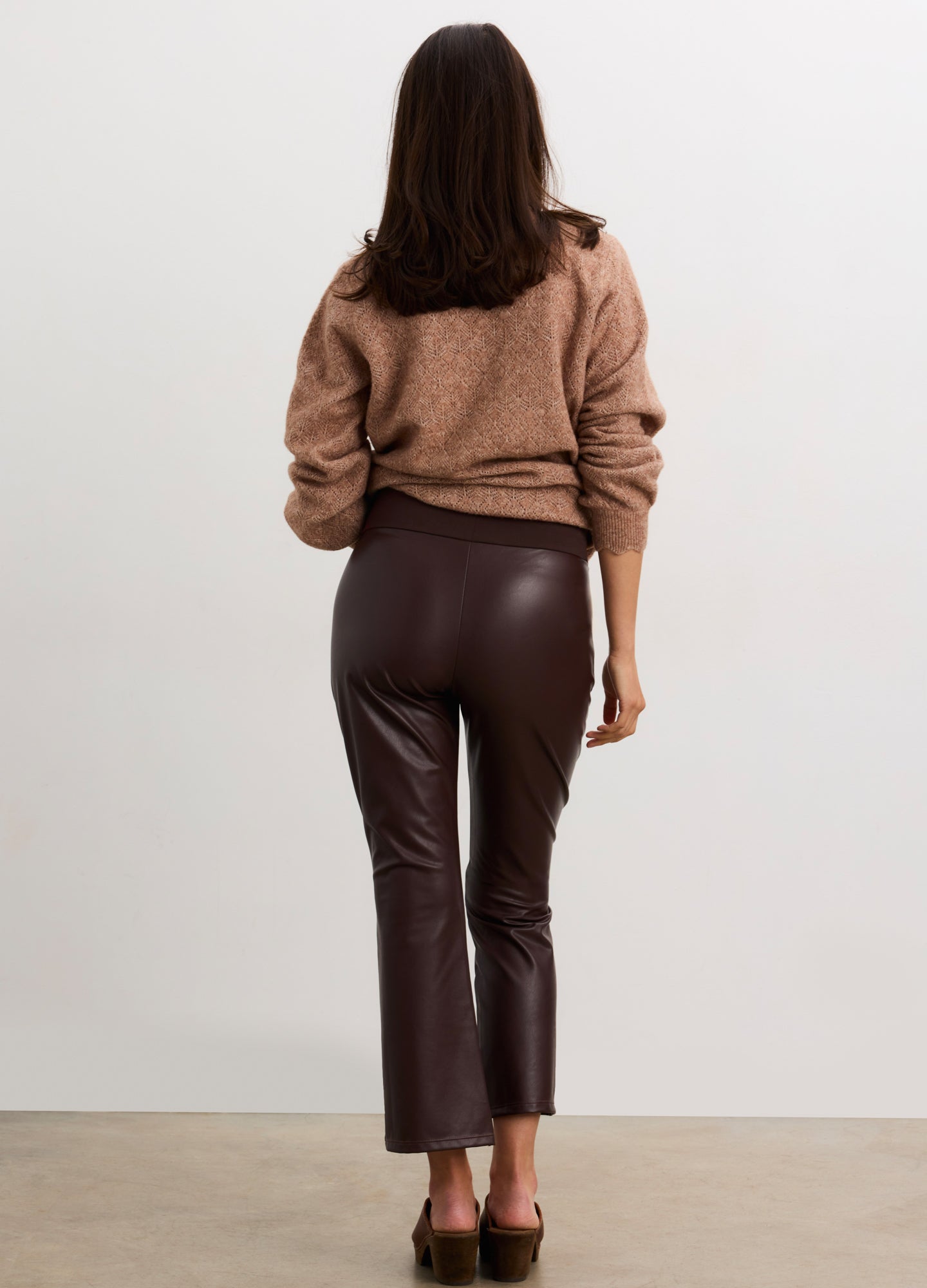 Pregnant woman wearing a brown sweater and leather pants against a white background