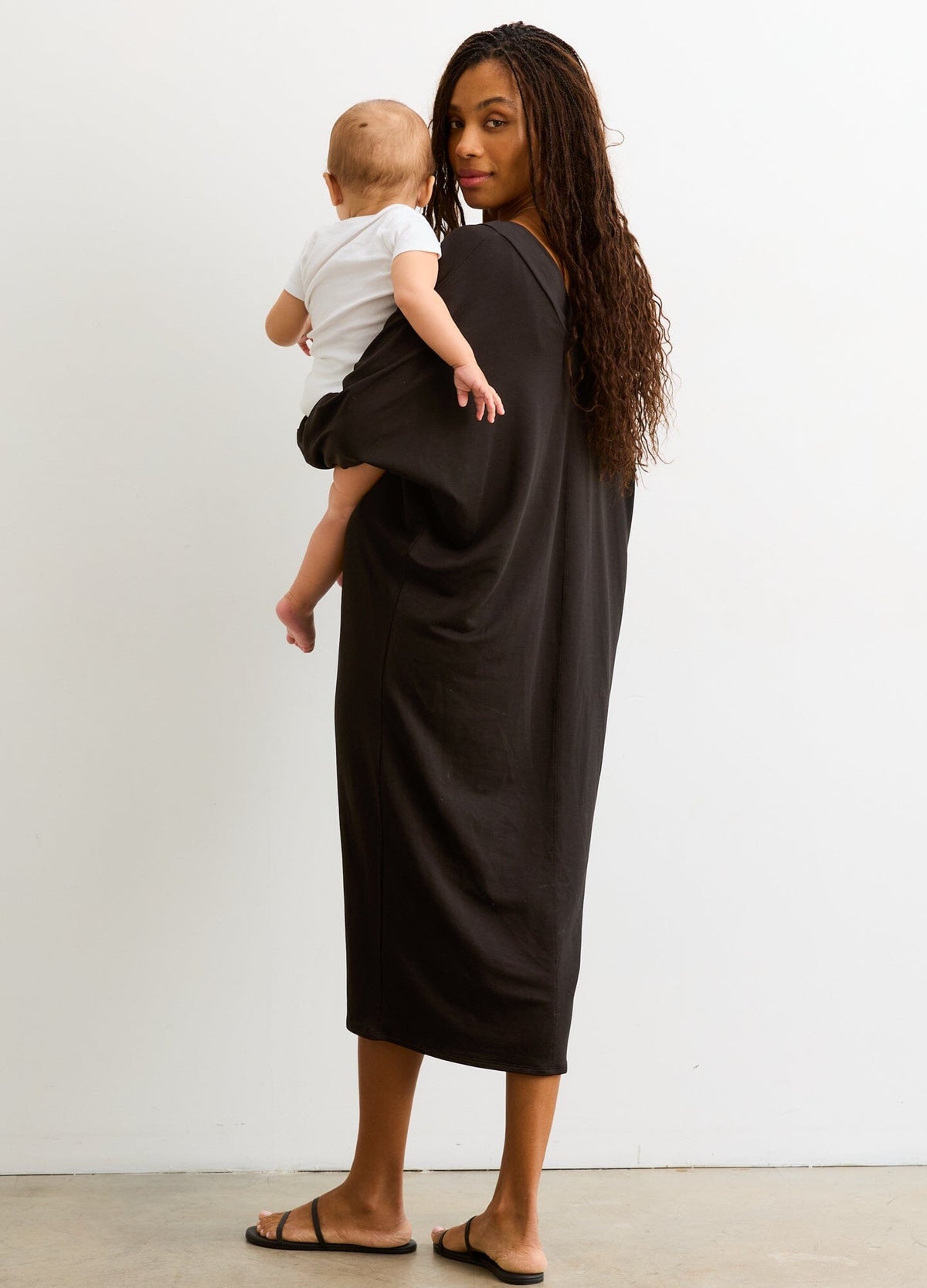 The Visitor Dress - Black maternity dress with long sleeves and V-neck, designed for pregnancy, nursing, and postpartum, modeled with baby.