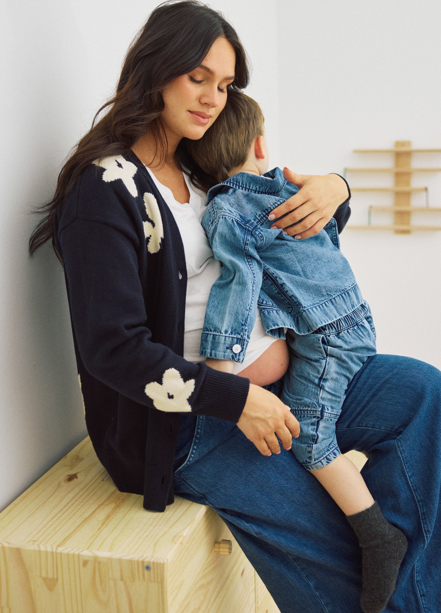 Navy blue cardigan with white flower appliqués worn over casual maternity jeans by a seated pregnant woman holding a toddler