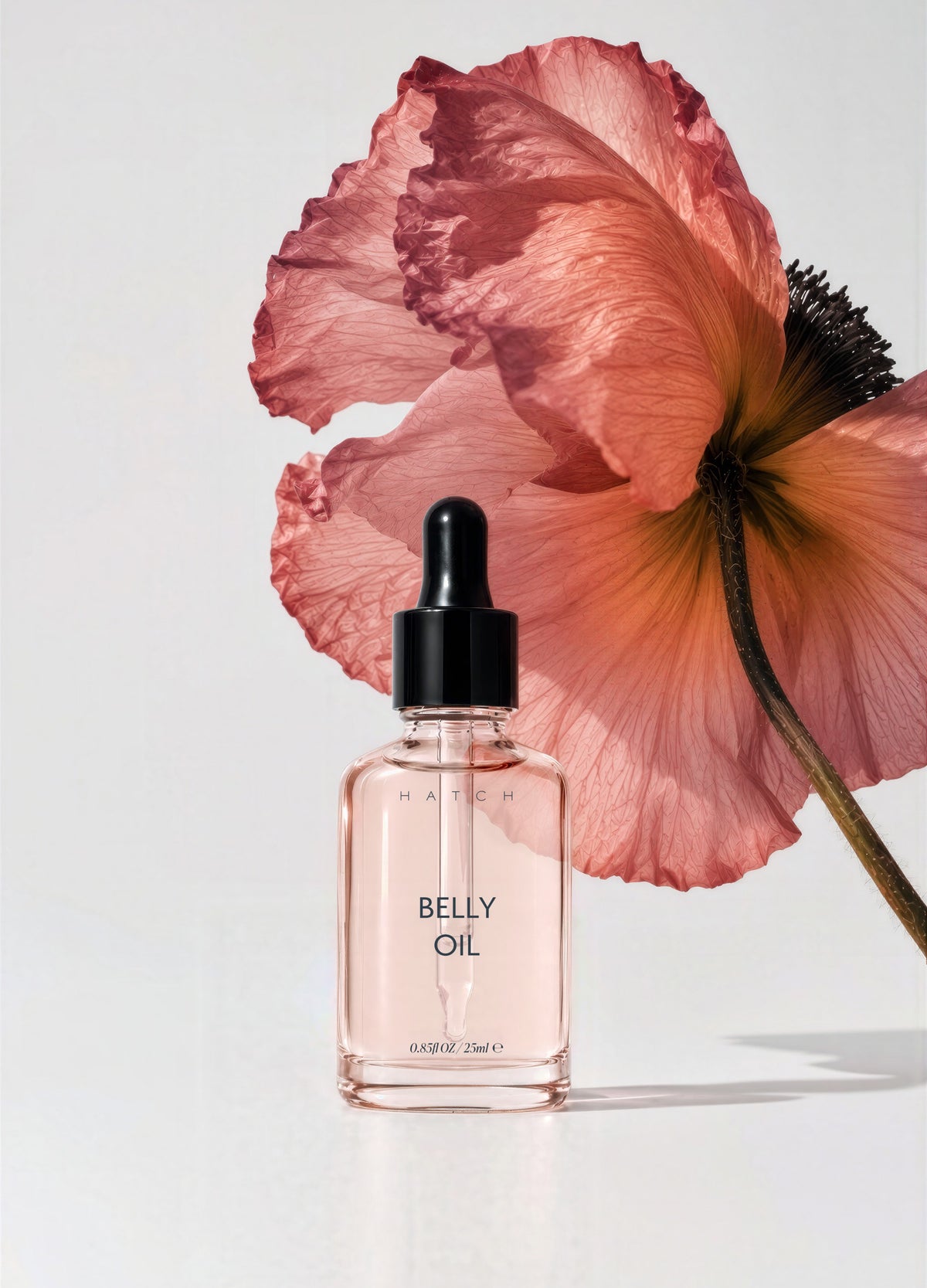 Clear glass dropper bottle with pale pink liquid labeled Mini Belly Oil, set against large pink flower background.
