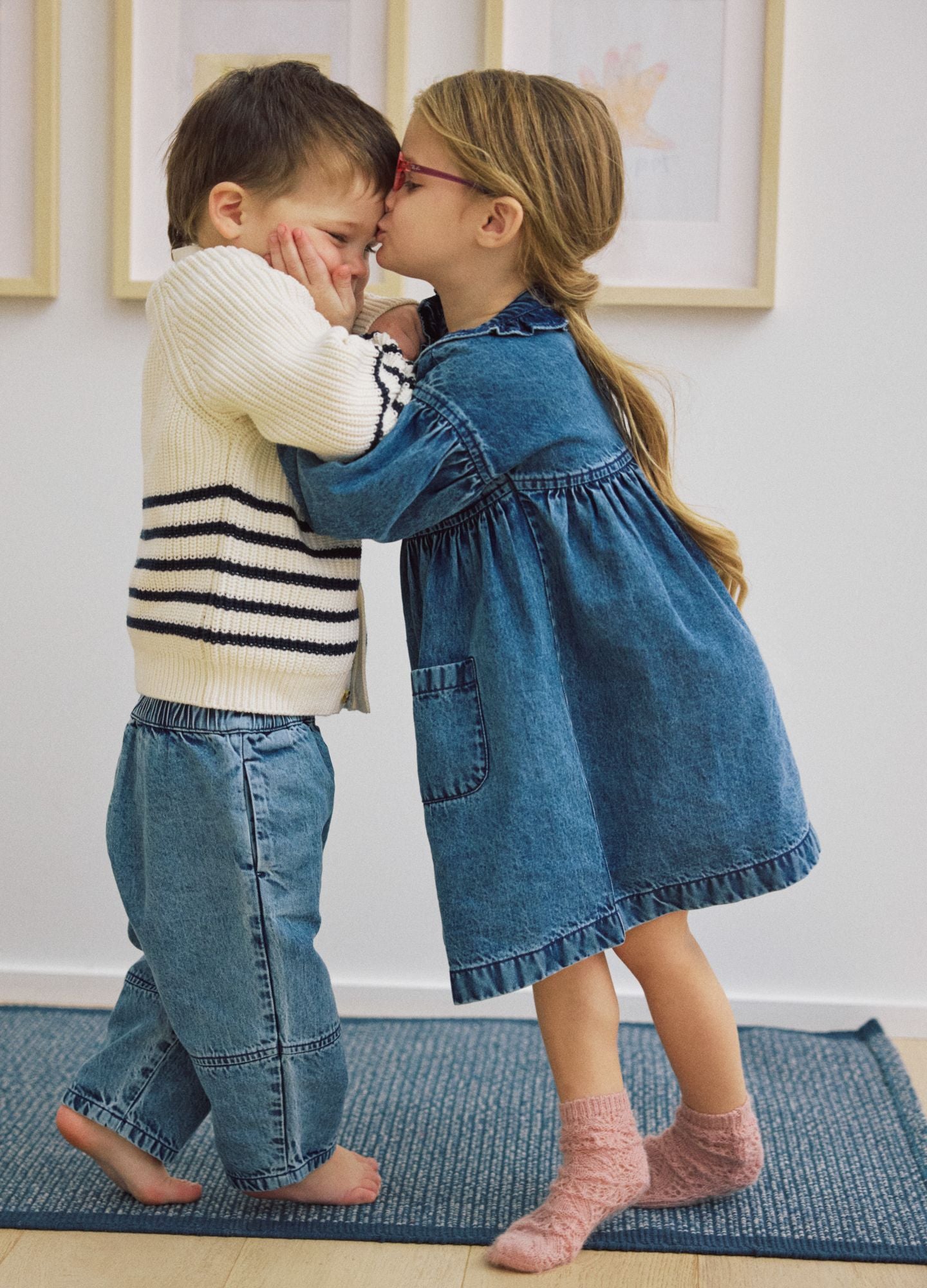 Blue denim dress with gathered waist and pockets worn by a young girl with glasses and pink socks, facing a boy in cream and